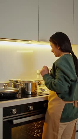 From-behind,-a-happy-brunette-girl-with-Brown-skin-in-a-green-sweater-and-a-beige-apron-prepares-food-using-a-saucepan-while-in-a-modern-kitchen-in-an-apartment-in-the-evening